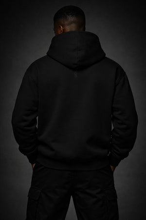 Back view of a black premium streetwear hoodie worn by a Black male model, showcasing the hood structure and a discreet tonal rear neck insignia in a monochrome studio setting.