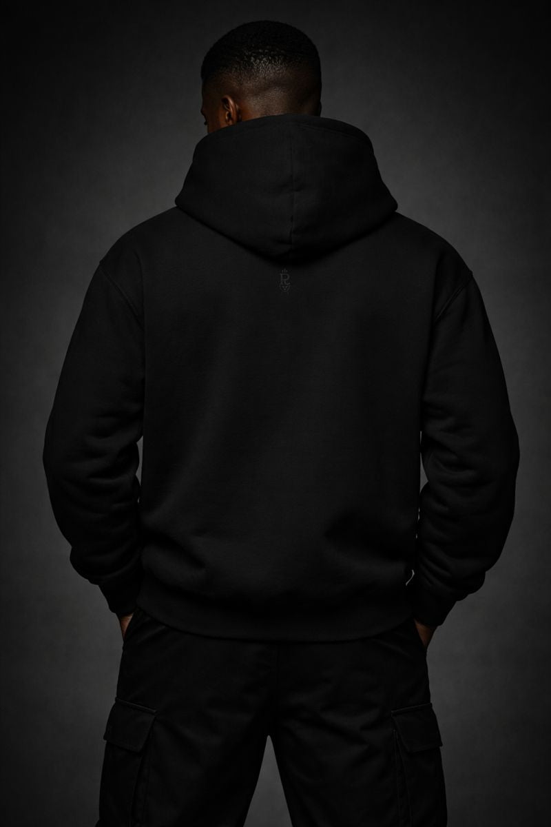 Front view of a black heavyweight streetwear hoodie worn by a Black male model, featuring a subtle tonal skull emblem on the chest and a minimalist black-on-black design against a dark studio background.