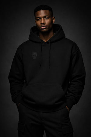 Front view of a black heavyweight streetwear hoodie worn by a Black male model, featuring a subtle tonal skull emblem on the chest and a minimalist black-on-black design against a dark studio background.