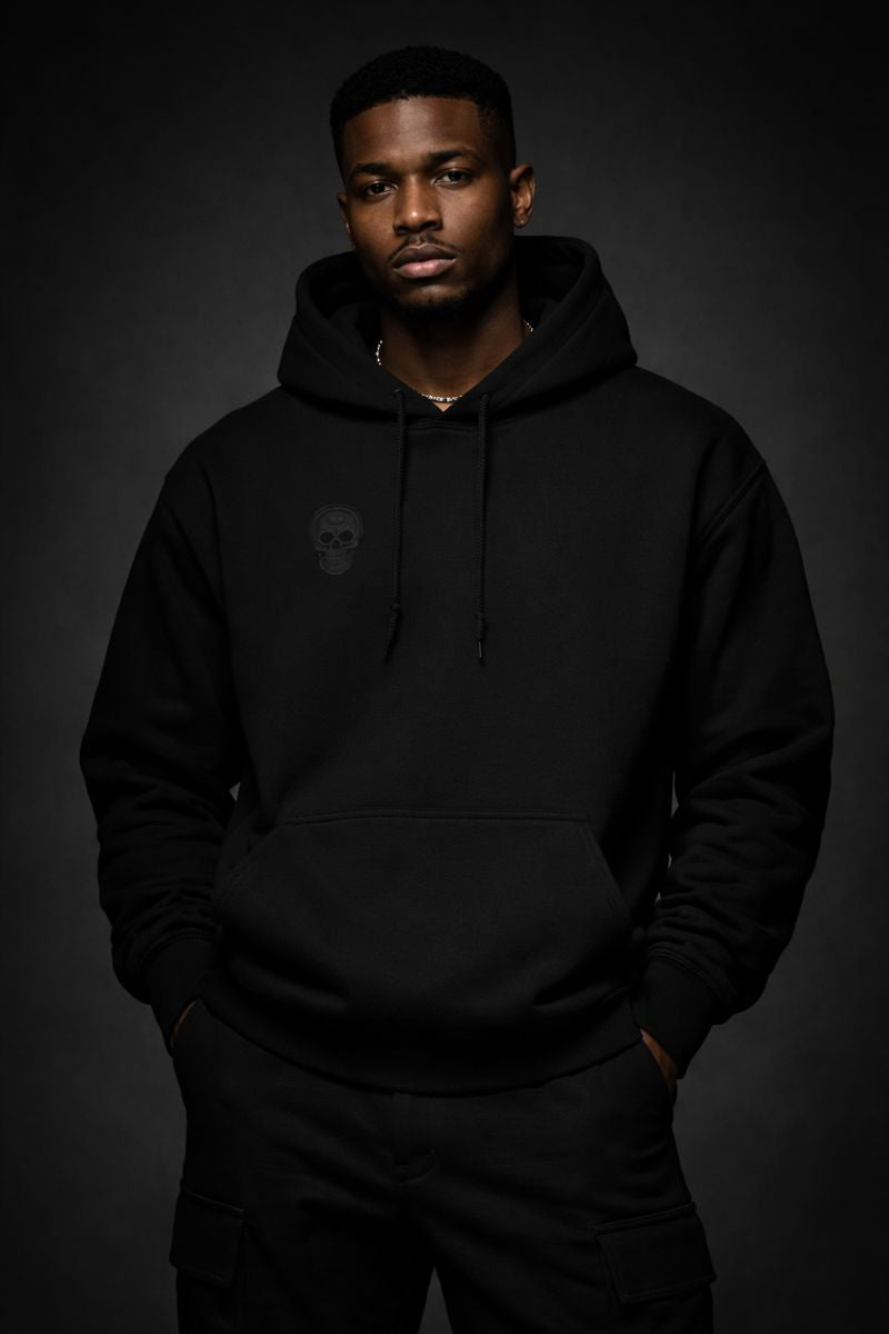 Front view of a black heavyweight streetwear hoodie worn by a Black male model, featuring a subtle tonal skull emblem on the chest and a minimalist black-on-black design against a dark studio background.