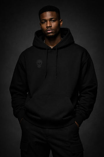 Front view of a black heavyweight streetwear hoodie worn by a Black male model, featuring a subtle tonal skull emblem on the chest and a minimalist black-on-black design against a dark studio background.