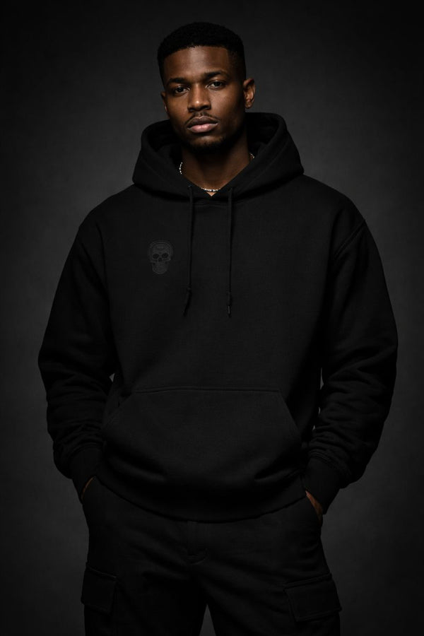 Front view of a black heavyweight streetwear hoodie worn by a Black male model, featuring a subtle tonal skull emblem on the chest and a minimalist black-on-black design against a dark studio background.