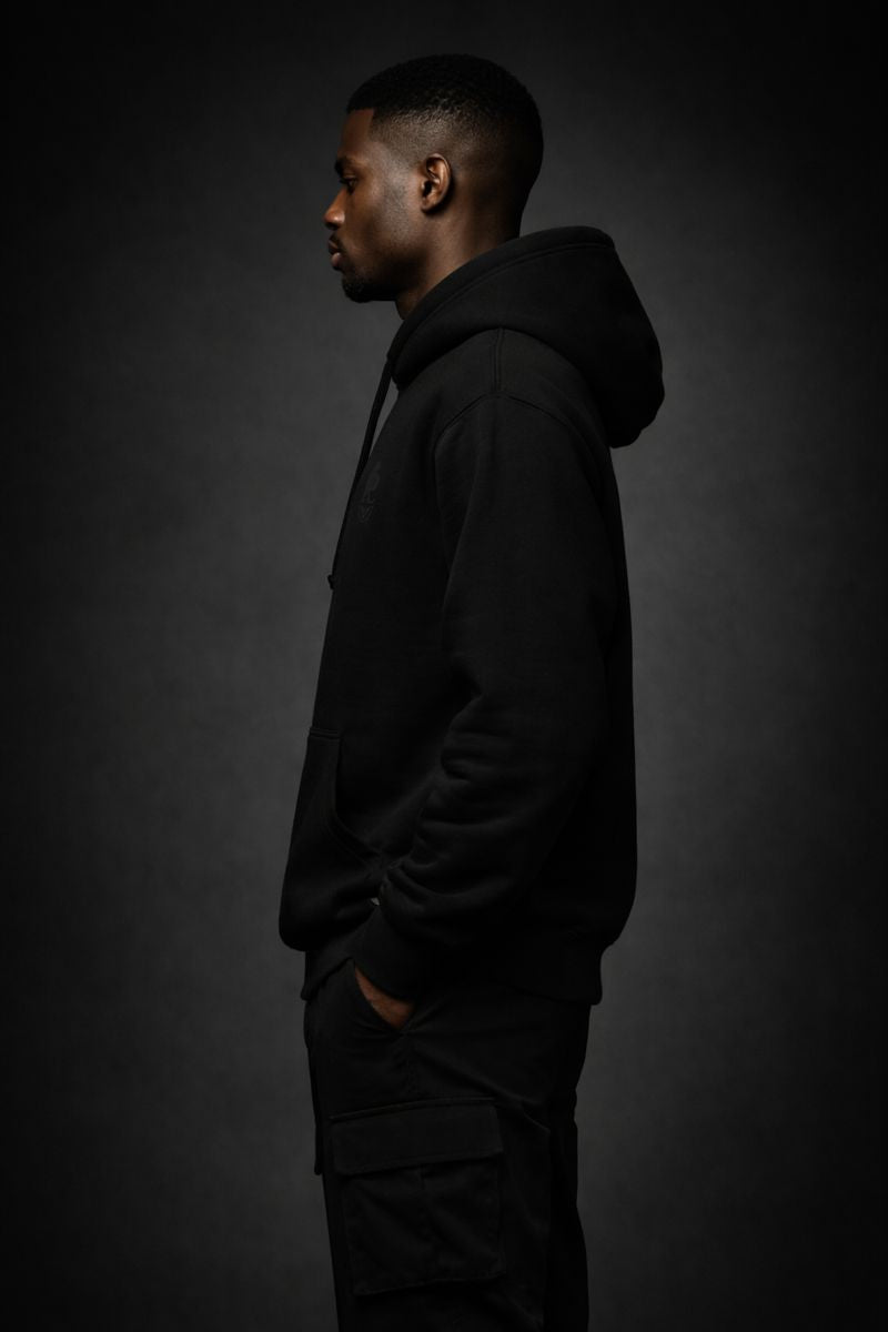 Side profile view of a black luxury streetwear hoodie worn by a Black male model, highlighting the hoodie’s silhouette, fabric weight, and clean minimalist construction in low-key lighting.