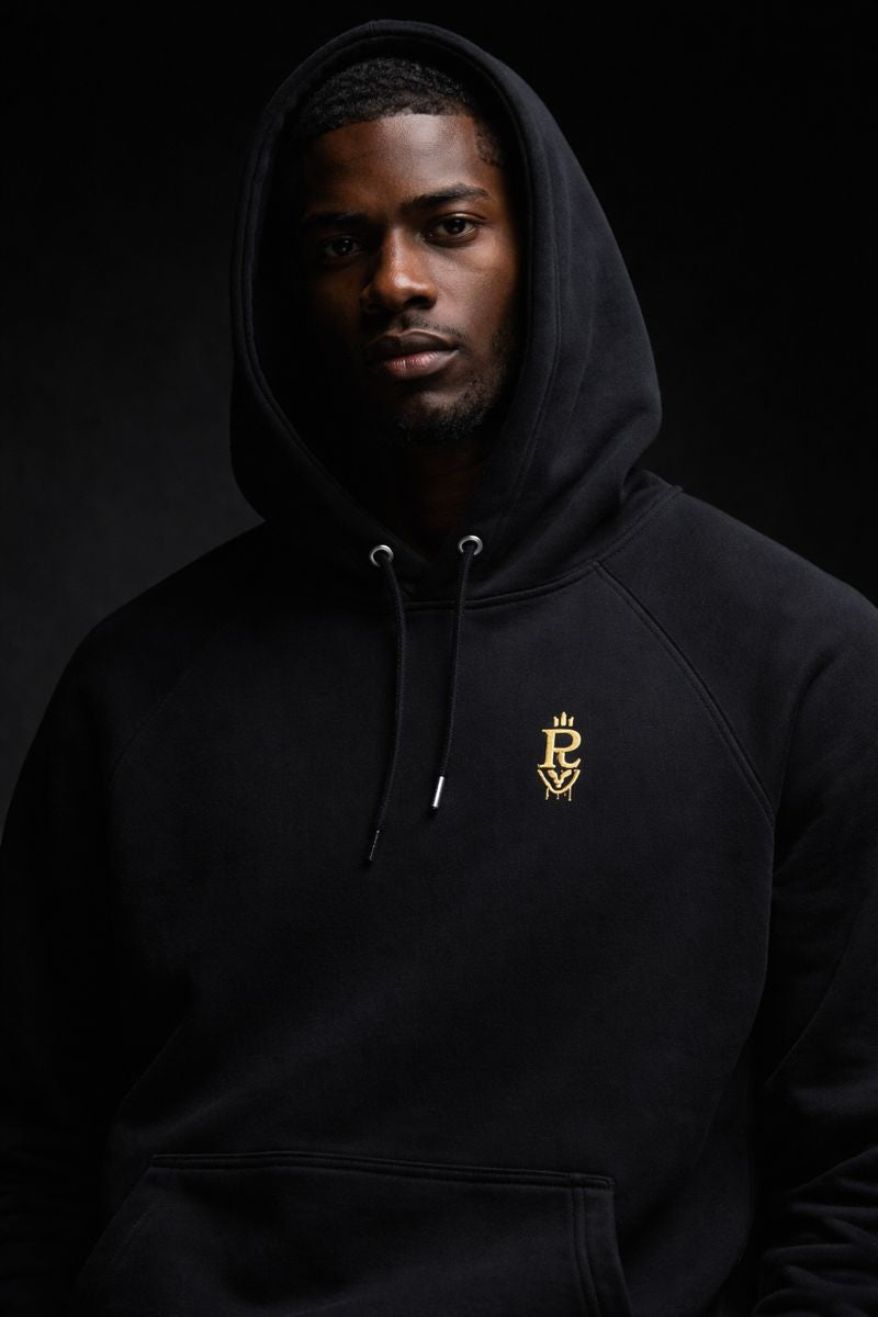 Black premium streetwear hoodie with gold chest crest and symbolic back graphic worn by a Black male model in a minimalist luxury photoshoot.